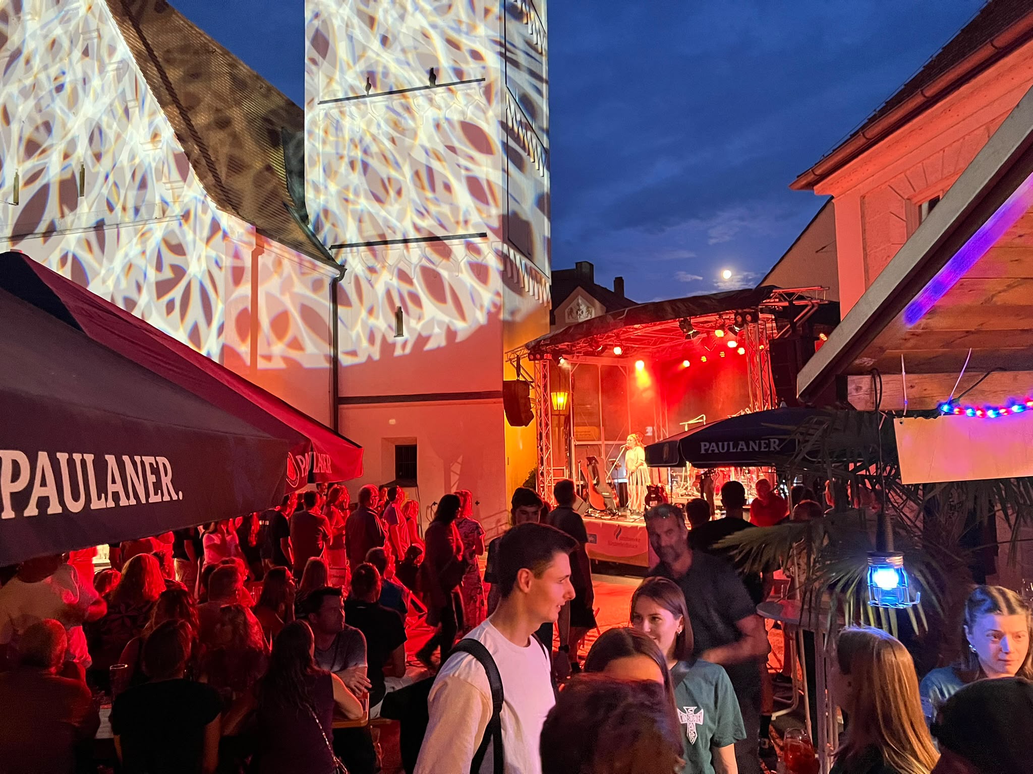 Nächtliche Bühnenshow mit rotem Bühnenlicht und Gobo-Projektion auf der Fassade, davor feiernde Menge
