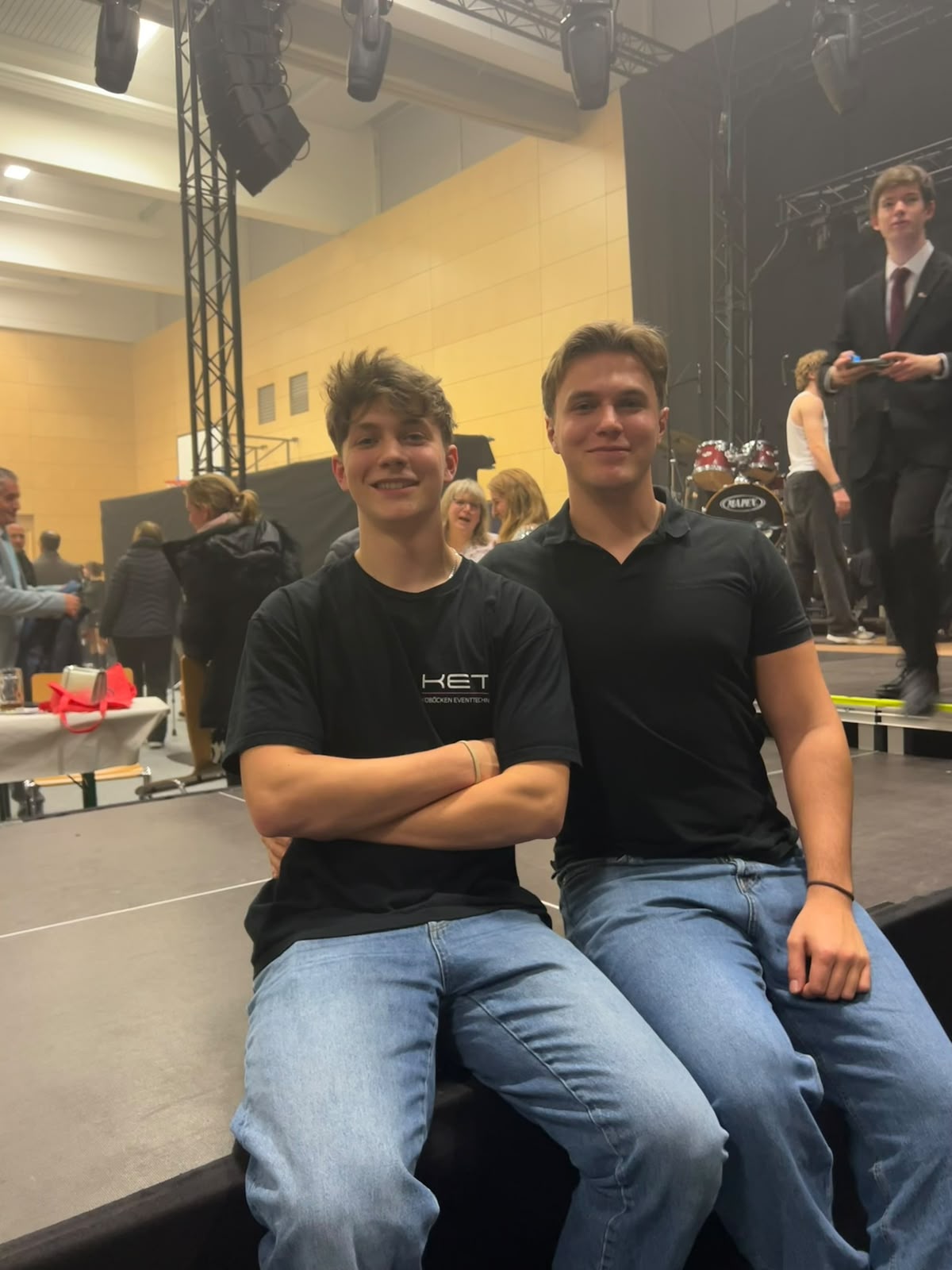 Nils und Lars Koböcken sitzen gemeinsam am Bühnenrand bei einer Show im Gymnasium Olching, einer der beiden im KET-Shirt